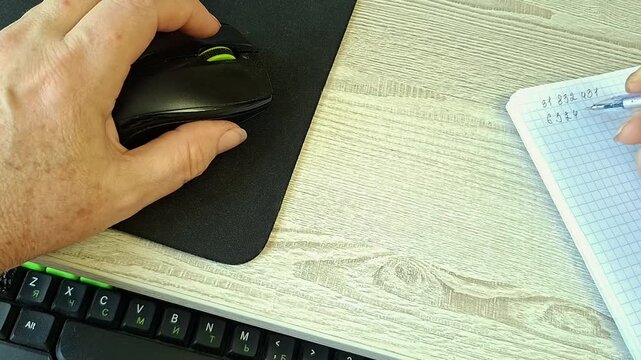 Senior person using computer mouse while writing numbers in notebook