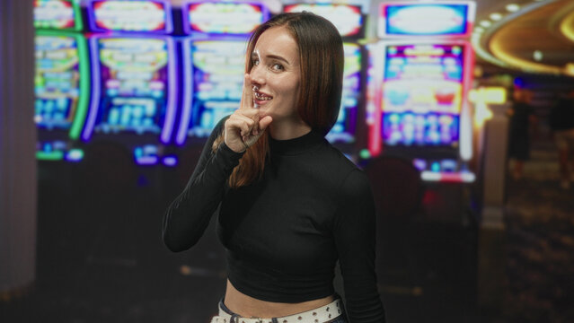 Woman redhead shushes with finger to lips beside slot machines in a gaming building, wearing black crop top and studded belt; playful secrecy.