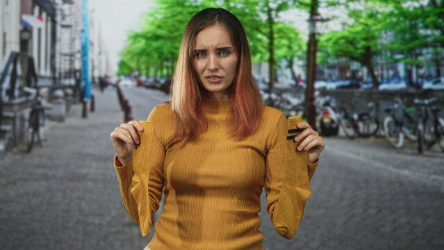 Woman holding a pair of yellow socks at shoulder height, frowning with knitted eyebrows and slightly hunched posture on a bike lined cobblestone street; frustration.