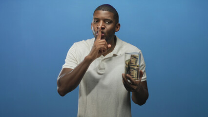 Young man holds glass jar of bundled dollar bills and presses finger to lips for silence in studio  secrecy. © Krakenimages.com
