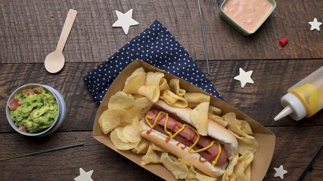 Panning from left to right over a wooden table with grilled hotdog topped with yellow mustard and red ketchup on a bun in paper tray with potato chips, various toppings, condiments, stars, and an Amer