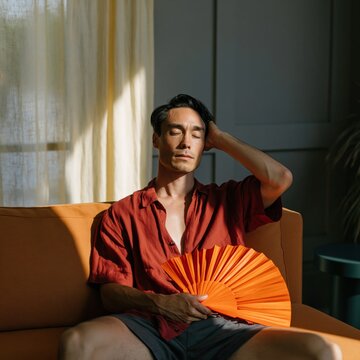 Man sitting on sofa fanning himself with orange hand fan in sunlight wearing red shirt for summer lifestyle and editorial background