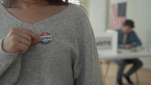 Woman shows voted sticker on sweater at voting building with flag and poll worker behind; patriotism civic duty.