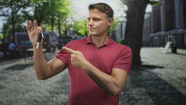 Man holding car keys and pointing index finger on cobbled street plaza with trees and benches in front of brick buildings wearing red polo shirt; confidence trust.