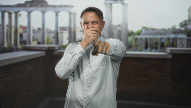 Man points finger at camera, laughs and alternately covers mouth while wearing a light patterned shirt on a building rooftop terrace with brick parapet and classical columns; playful confidence.