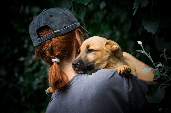 An intimate back-view portrait of a young woman with a baseball cap holding a light brown puppy in her arms.