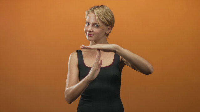 Blond woman forms timeout gesture with hands and raised palms while wearing black tank top in studio under warm lighting; calm pause.