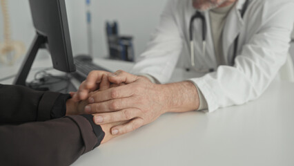 Fototapeta premium Doctor holds patient's hands on desk, white coat and stethoscope visible in clinic building; compassion support reassurance.