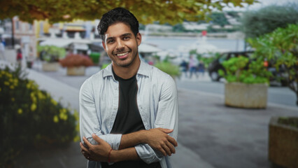 Obraz premium Young man with arms crossed hugging his chest on a waterfront street promenade, smiling in casual shirt and bracelet; calm.