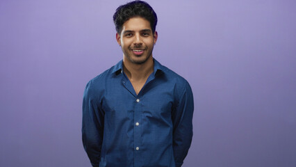 Obraz premium Young man standing with hands behind back in studio, wearing a blue button shirt against a purple backdrop, eyes moving from closed to open with a slight smile; calm confidence.