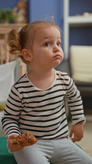 Fototapeta premium Little blonde girl in a striped shirt holding a pastry with one hand, seated and looking sideways in a home building; quiet curiosity.