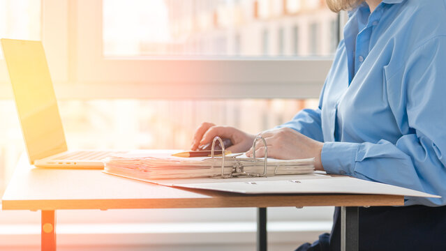 Professional person types on laptop at wooden desk. Warm sunlight streams through large window. City buildings visible outside. Focused demeanor suggests dedication