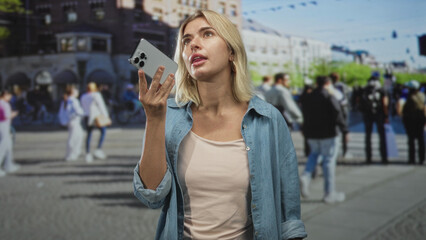 Fototapeta premium Woman holds smartphone to mouth, speaks into it on a busy street near a blurred crowd and building facade; urban communication concern.