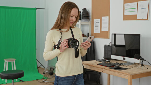Woman photographer holds dslr camera and raises smartphone for selfie in a studio with green screen and computer monitor; focused workflow.