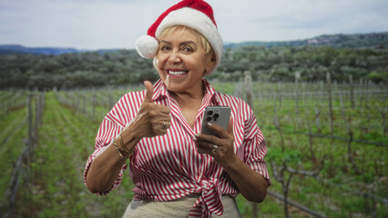 Obraz premium Woman wearing santa hat gives thumbs up while holding smartphone in a vineyard forest; holiday cheer.
