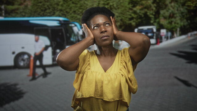 Woman covers ears with both hands beside parked coach buses on a street shuttle pickup, wearing yellow blouse and squinting in daylight; travel noise stress.