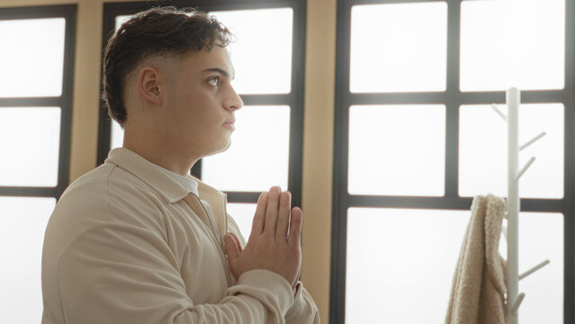 Young man palms together in a prayer gesture, hands pressed at chest in a bright yoga studio; serenity focus.