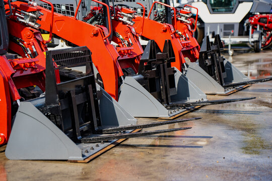 A row of new red skid steer loaders equipped with industrial pallet forks parked in a line at a dealership lot ready for construction or farming work