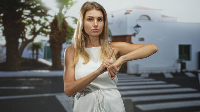 Woman in a white dress points to her wrist and mimes checking time at a crosswalk on street in front of a white building; impatience waiting.