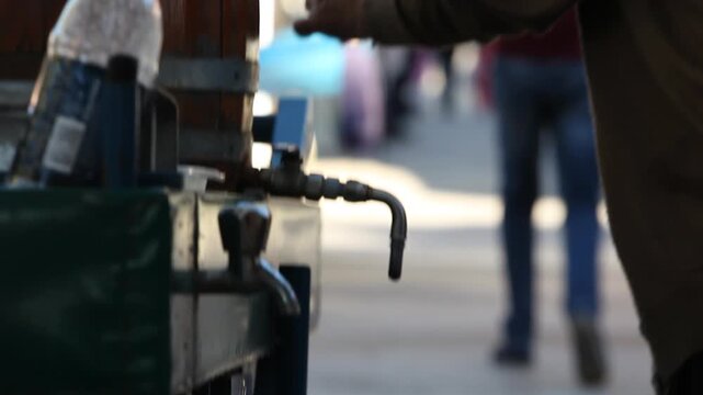 Close-up of a street vendor pouring a traditional red beverage from a wooden barrel into a glass.