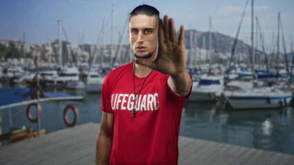 Fototapeta premium Man lifeguard in red shirt with whistle raising hand palm forward to stop on marina dock pier; vigilance duty.
