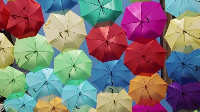 A dense arrangement of multi-colored umbrellas creates a festive overhead canopy. Sunlight filters through the fabric panels, highlighting saturated hues and soft shadows in a joyful urban installatio