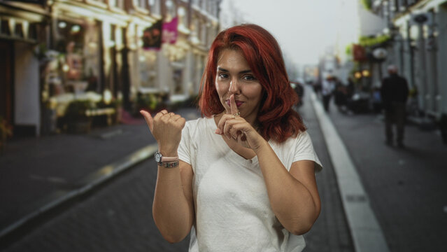 Woman with index finger to lips and thumb pointing sideways on a street, red hair and white t shirt visible; playful secrecy.
