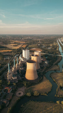 Industrial power plant with cooling towers surrounded by green fields and a canal under a clear blue sky, captured at sunset &mdash; symbol of modern energy and landscape harmony.