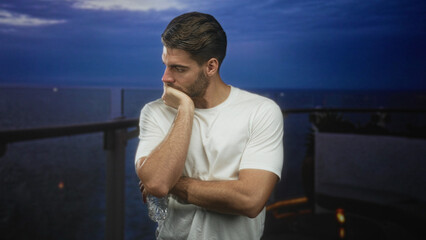 Fototapeta premium Man with beard in white shirt holding water bottle, hand on chin leaning on balcony railing of building overlooking ocean at dusk; quiet contemplation.