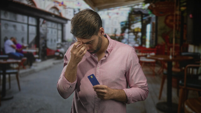Hispanic man with beard wearing pink shirt holding creditcard pinches nose bridge on street cafe terrace amid empty tables; financial anxiety.