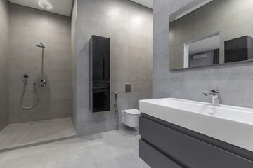 A bathroom features a large shower with a water fixture, a sink with a faucet, and a toilet. Gray tiles cover the walls and floor. The space has clean lines and a simple design © Alhim