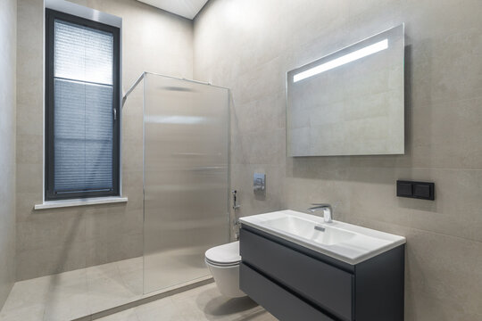 A bathroom with a glass shower, a toilet, and a sink with a mirror. Light enters from a window. The tiles on the walls and floor are clean and simple. The design is functional and tidy