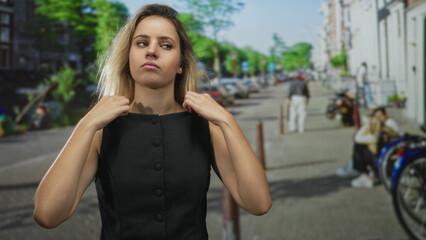 Fototapeta premium Woman young blonde adjusts dress collar with both hands while standing near parked bicycles on a busy city street; defiance attitude.