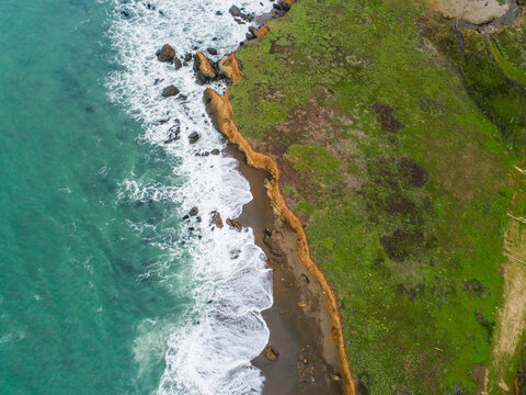 Aerial view of the rugged coastline near Boucher Trailhead North, featuring turquoise waves crashing against rocky cliffs and green coastal bluffs in San Simeon, California, United States.