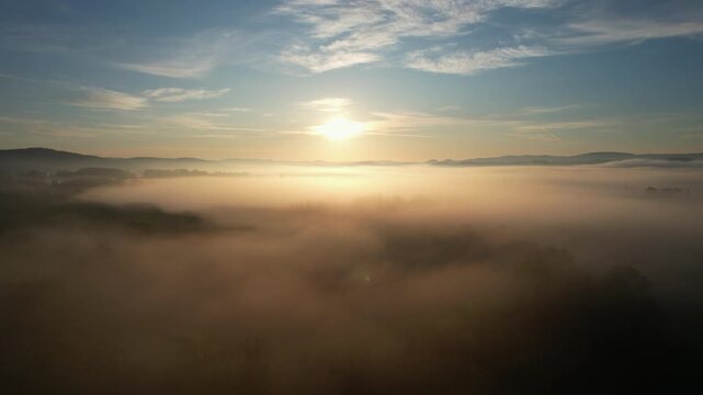 Cinematic drone flight ascending through thick white morning mist directly towards a glowing sun on the horizon. Dreamy sunrise panorama over a sea of clouds and distant mountain ridge, Lower Silesia.