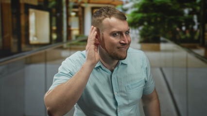Fototapeta premium Man with hand cupped to ear leaning forward on indoor building walkway near glass railing and plants; curious listening communication.