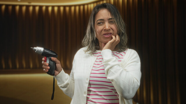 Middle-aged hispanic woman holding a cordless screwdriver, hand pressed to cheek and wincing against studio curtains and warm stage lighting; discomfort.