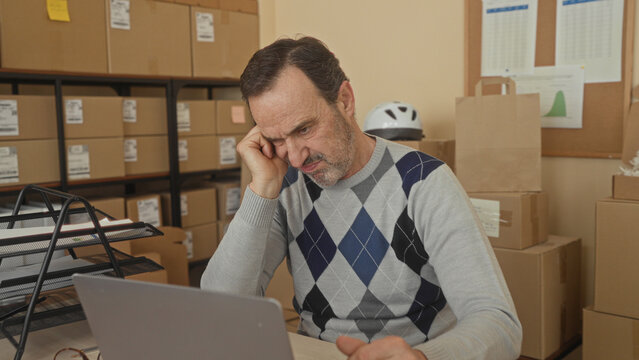 Middle aged man at laptop with hand on cheek among stacked cardboard boxes in a building; small business stress.