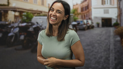 Fototapeta premium Woman laughing with hands clasped over her stomach on a street with parked motorcycles and cobblestone; joy warmth.
