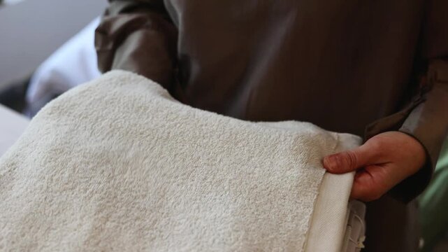 Person hands adjust folded cream towels and linens on white surface beside blurred shelf storage, creating neat domestic organization scene with warm light, soft textures, muted colors, and calm