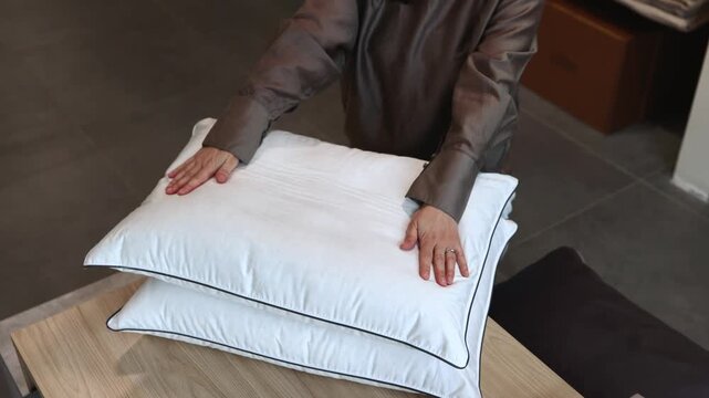 Cropped person in satin sleepwear adjusts stacked white pillows on light wood surface inside modern home interior, with soft warm light, dark tiled floor, blurred background, smooth fabric texture