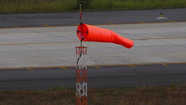 Orange windsock blowing in the wind next to airport runway tarmac, showing wind direction on cloudy day, static medium shot