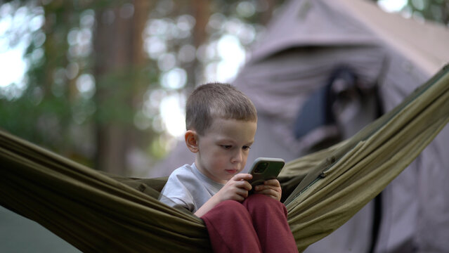 Little boy relaxes in green hammock using phone at campsite. 