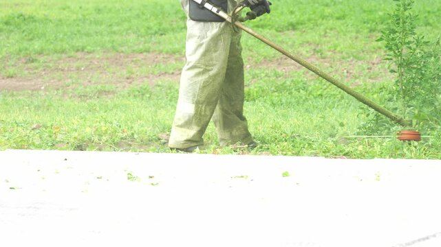Green grass stretches beneath a lawnmower. Worker wears gloves and work pants. Background shows grassy field with dirt patches. Ideal for landscaping or garden maintenance projects