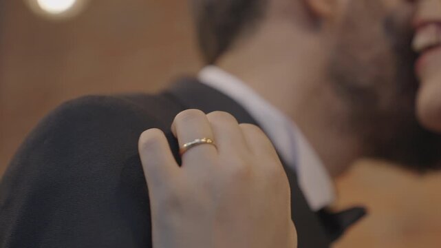 Close-up of a woman's hand wearing a gold wedding ring resting affectionately on a man's suit shoulder. The smiling, romantic couple is out of focus in the background.
