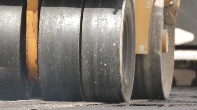Worn steel wheels press asphalt. Heavy machinery moves slowly. Smooth asphalt shows recent compaction. Construction site work in progress. Industrial scene for roadwork projects