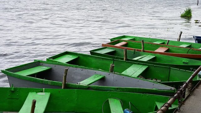 A row of empty green rowboats gently bobs on the choppy water of a lake, moored securely to a dock 