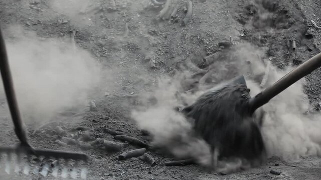 Detail shot of manual laborers using a shovel and rake to sort and move black charcoal and ash, creating thick gray dust. Ideal for traditional labor and rural industry themes.