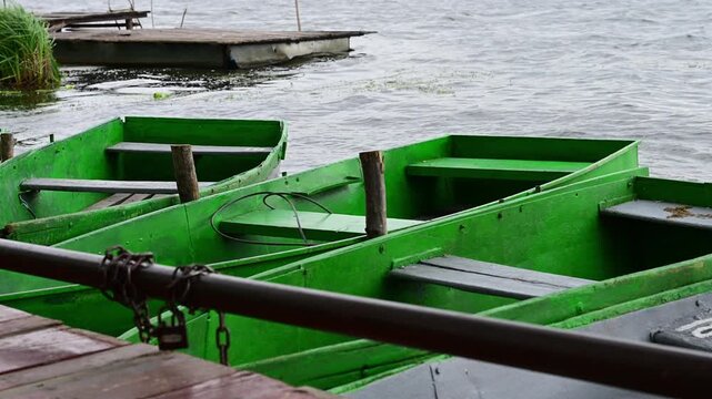 A row of empty green rowboats gently bobs on the choppy water of a lake, moored securely to a dock 