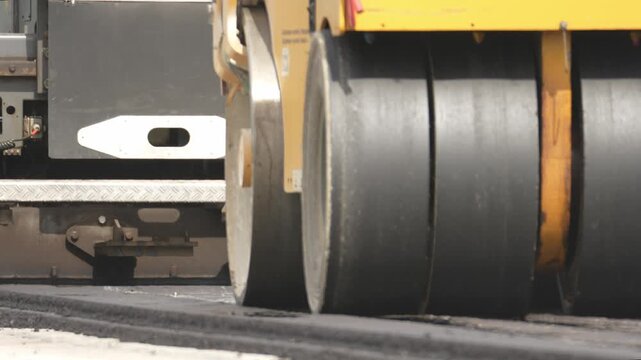 Close-up of a road roller's drums compacts fresh asphalt. Black cylindrical drums press down on the new road surface. Yellow machine body visible behind the rollers